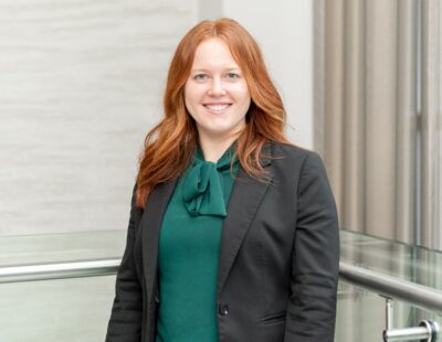 Kirstie Orton head shot image, wearing a green blouse and dark grey suit jacket, with auburn hair.